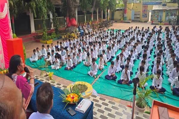 विद्यालय परिसर में हरी चटाइयों पर पंक्तियों में बैठे छात्र और मंच से माइक पर संबोधित करती महिला