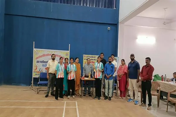 इनडोर बैडमिंटन कोर्ट पर पुरुष और महिला खिलाड़ी तथा प्राध्यापक एक पंक्ति में खड़े हैं, बीच में रखी मेज़ पर ट्रॉफी और सामने प्रतियोगिता का बैनर लगा है।
