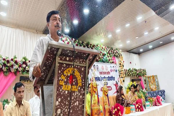 मंच पर लगे लकड़ी के पोडियम पर खड़े एक व्यक्ति माइक पर संबोधित करते हुए; पीछे फूलों की सजावट, प्रतियोगिता का बैनर और बैठे हुए अतिथि दिखाई दे रहे हैं।